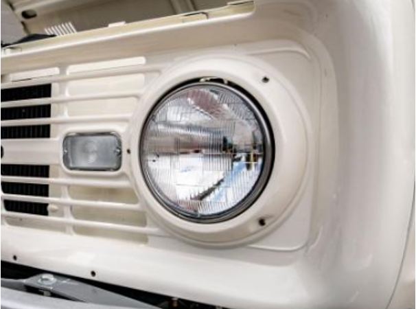 Close-up of OEM eyebrow grille on a 1966 Ford Bronco showing the scalloped sheet metal stamping above the round headlight and marker light in factory uncut white finish.